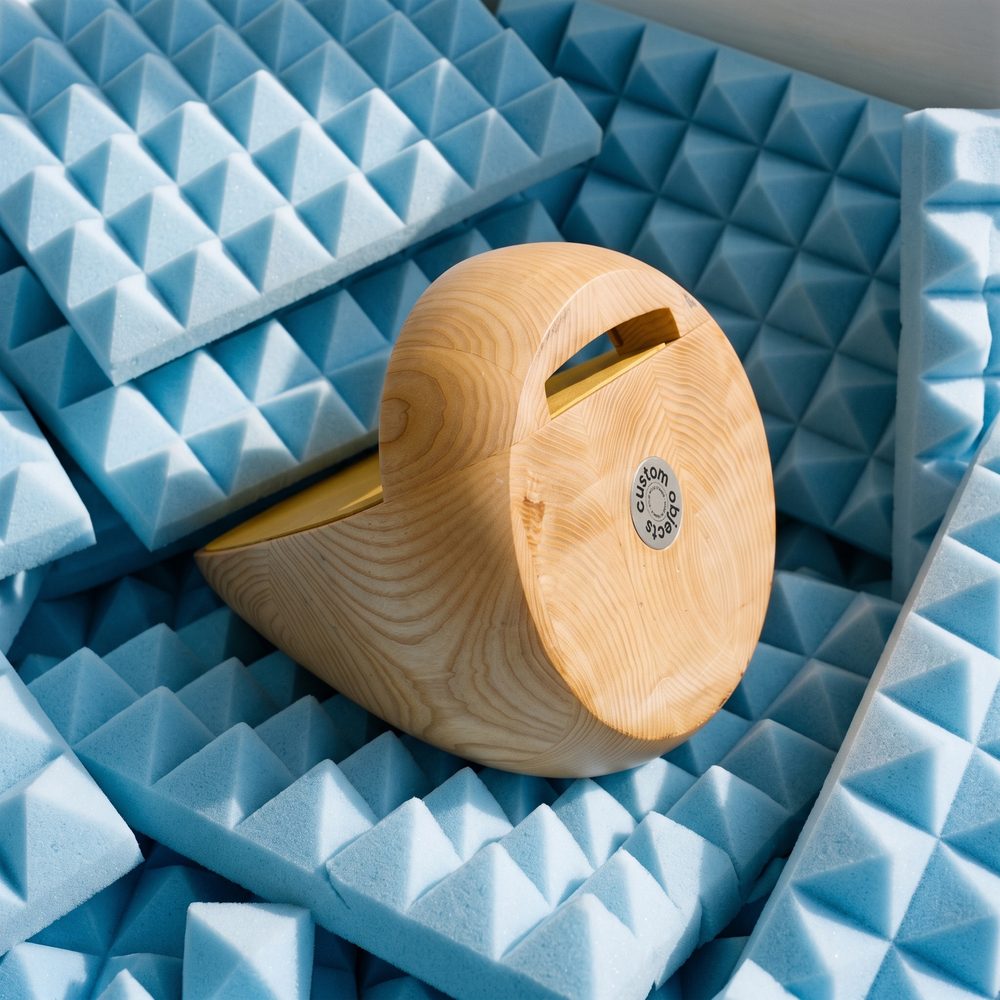 Stèle en bois clair couchée sur de la mousse acoustique bleue, avec le médaillon Custom Objects